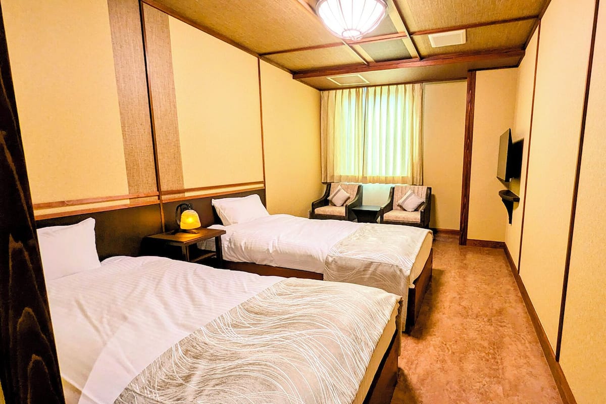Una habitación de hotel con dos camas, una mesita de noche con lámpara, una pequeña mesa y un televisor en la pared.