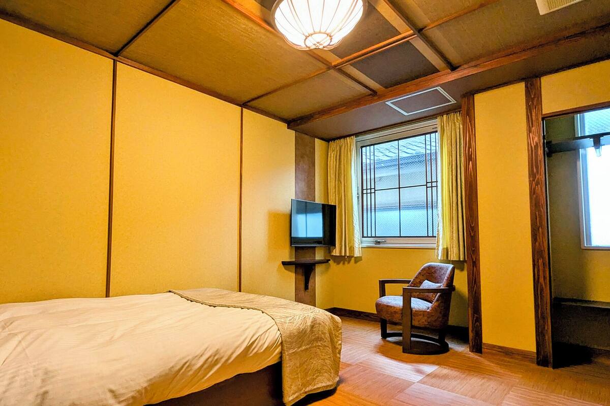 Una habitación con cama, silla, televisión y ventana con cortinas.