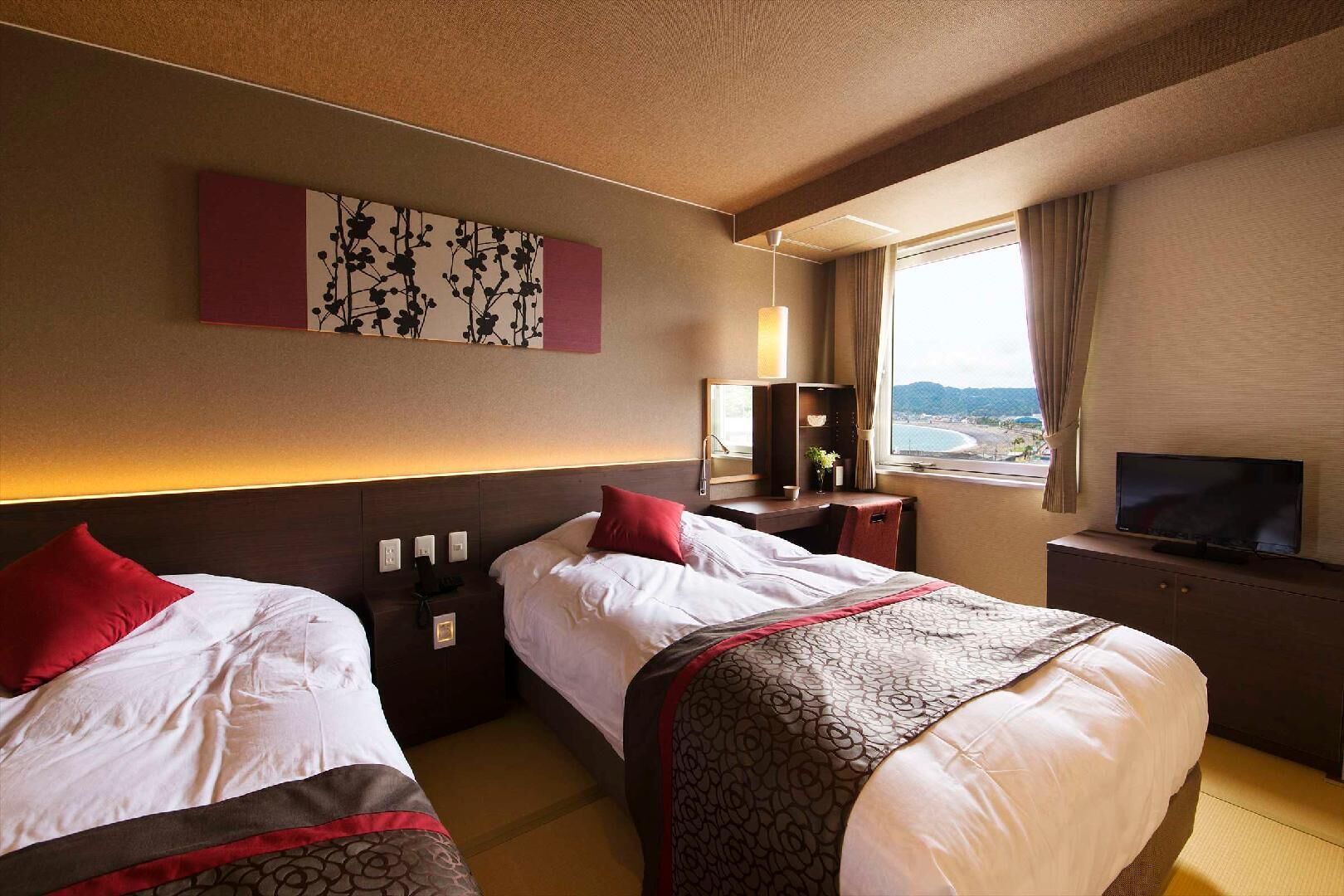 A hotel room with two beds, a TV, a desk with a lamp, a window with a view, and artwork on the wall.