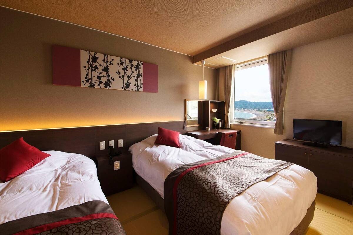 Een hotelkamer met twee bedden, een televisie, een bureau met lamp, een raam met uitzicht en kunst aan de muur.