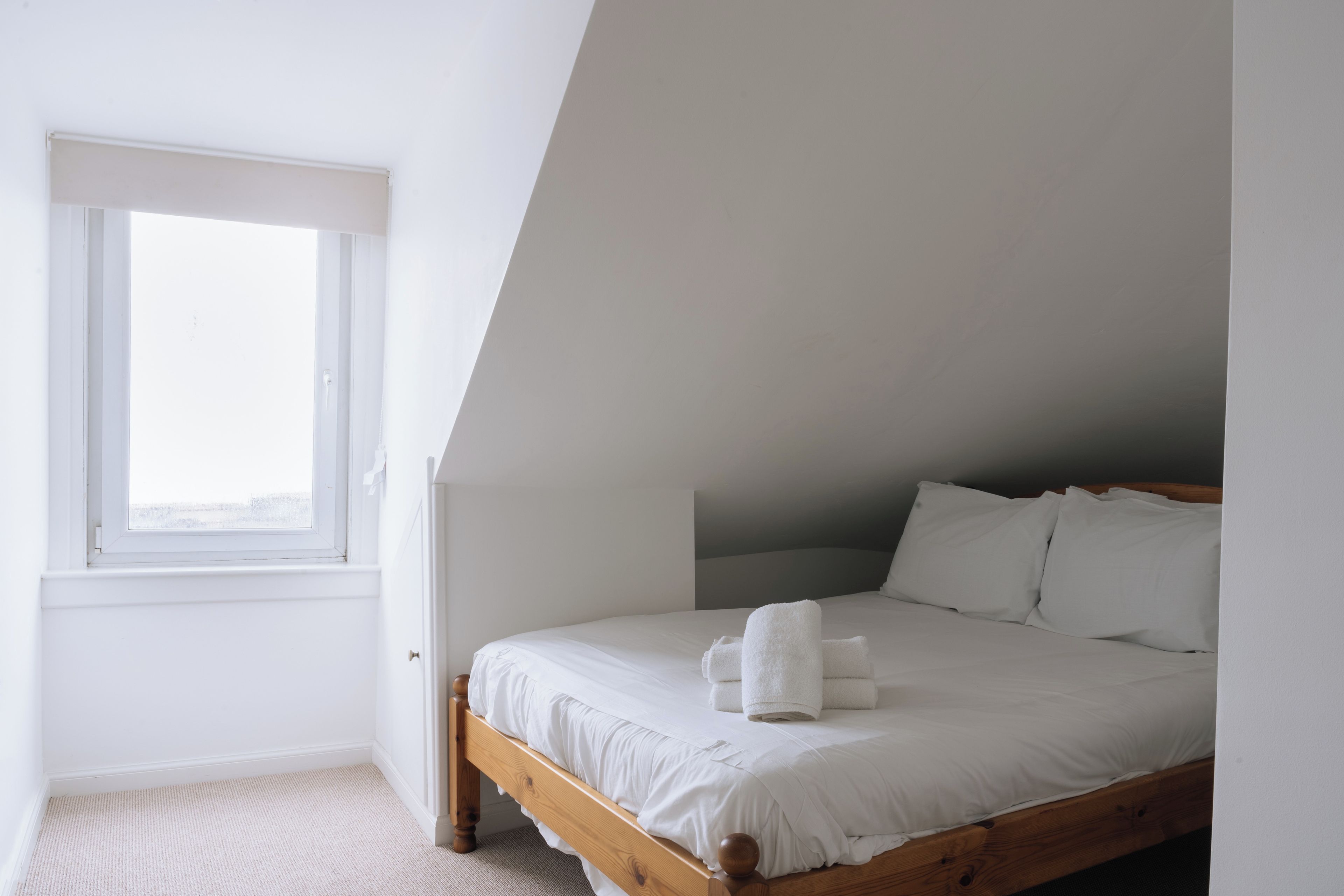 A bedroom with a sloped ceiling, a single bed with white bedding, and a window.