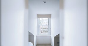 Corredor estreito com uma janela no final, que leva a um quarto com uma porta à vista.