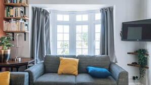 Ein Wohnzimmer mit grauem Sofa, einem gelben und einem blauen Kissen, einer Bücherwand, einem Fernseher und einer Pflanze.
