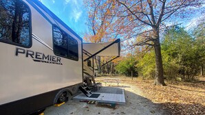 Un modèle Premier Ultralite, un type de véhicule récréatif léger, est stationné dans un terrain de camping, avec un patio couvert, des sièges d’extérieur et une table de pique-nique.
