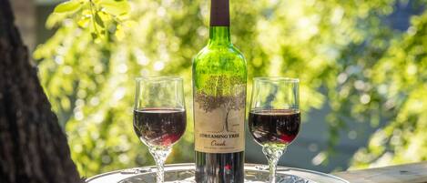 Una botella de vino tinto con dos copas sobre una bandeja, colocada al aire libre con un árbol y follaje de fondo.