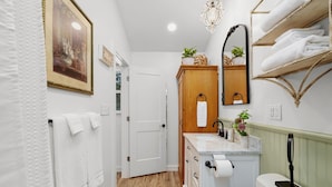 A narrow hallway with a wooden door, a white towel rack, a mirror, and a shelf with folded towels.