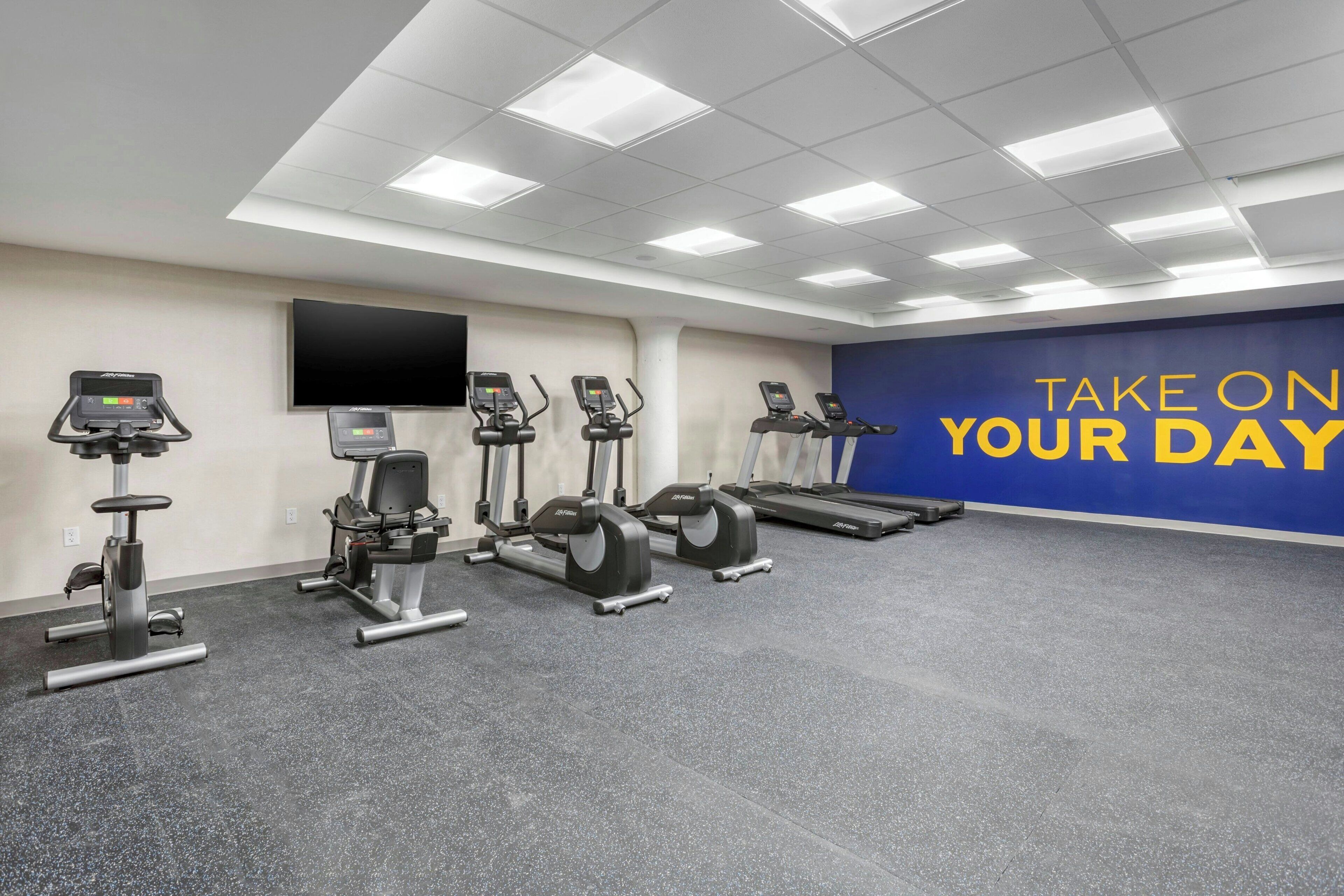 Een fitnessruimte met diverse trainingsapparaten, een groot scherm en motiverende tekst op de muur.