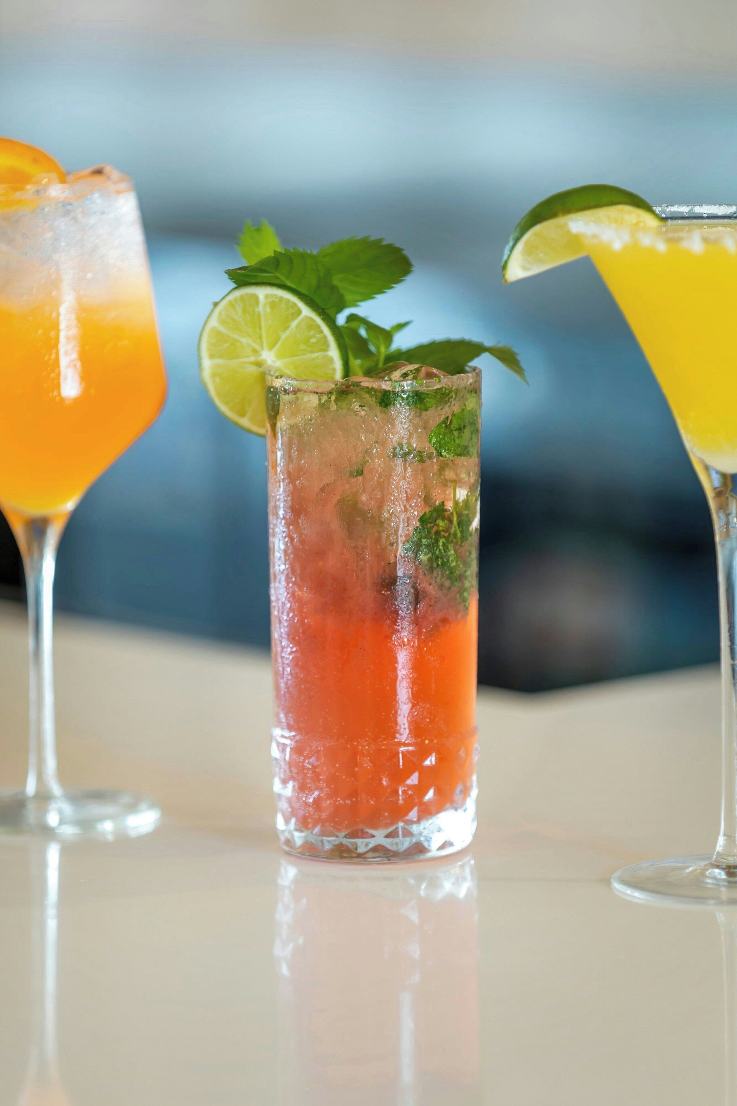 Three glasses of cocktails with ice and garnishes on a reflective surface.