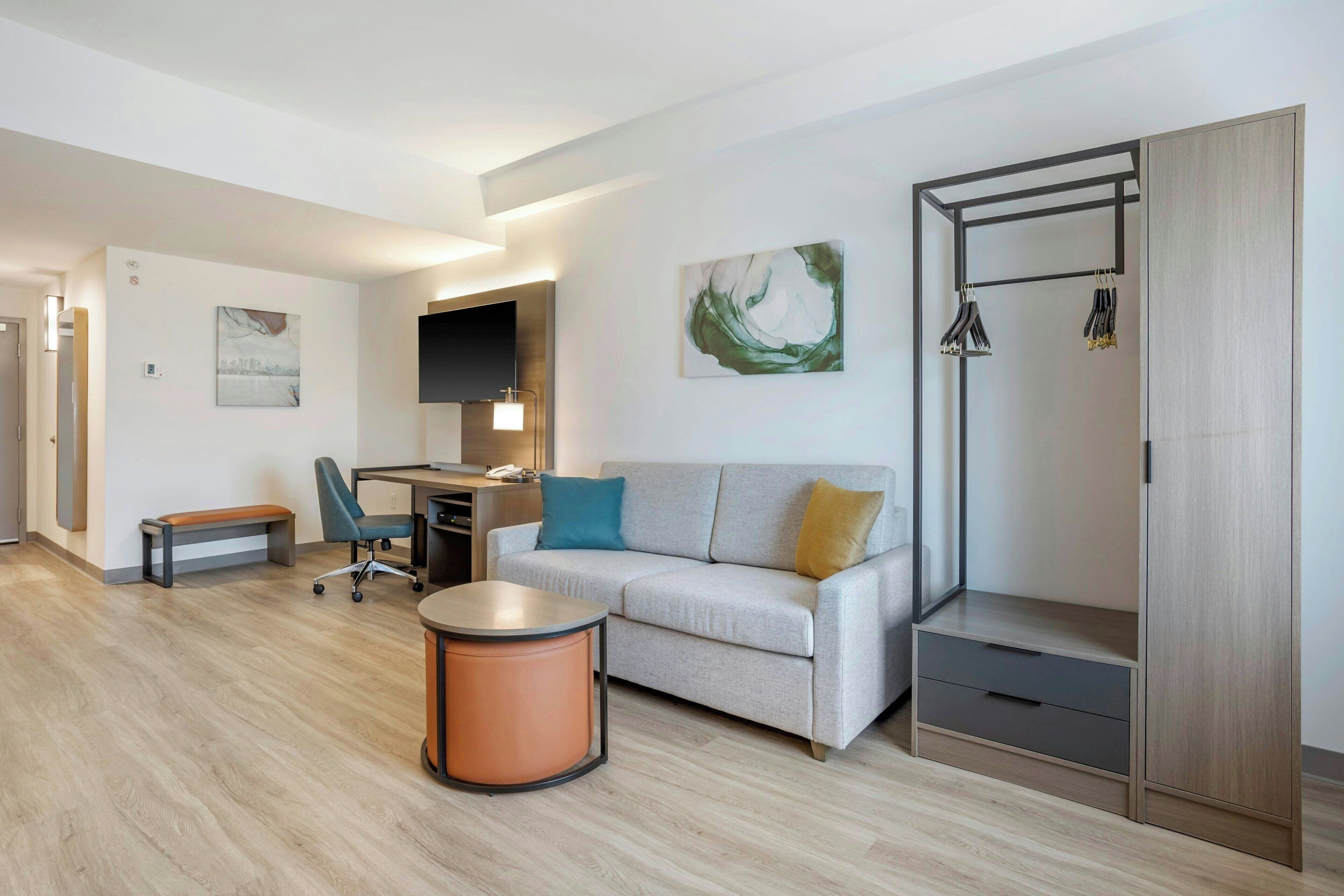 A modern hotel room with a sofa, a desk, a chair, a round table, and a wardrobe.
