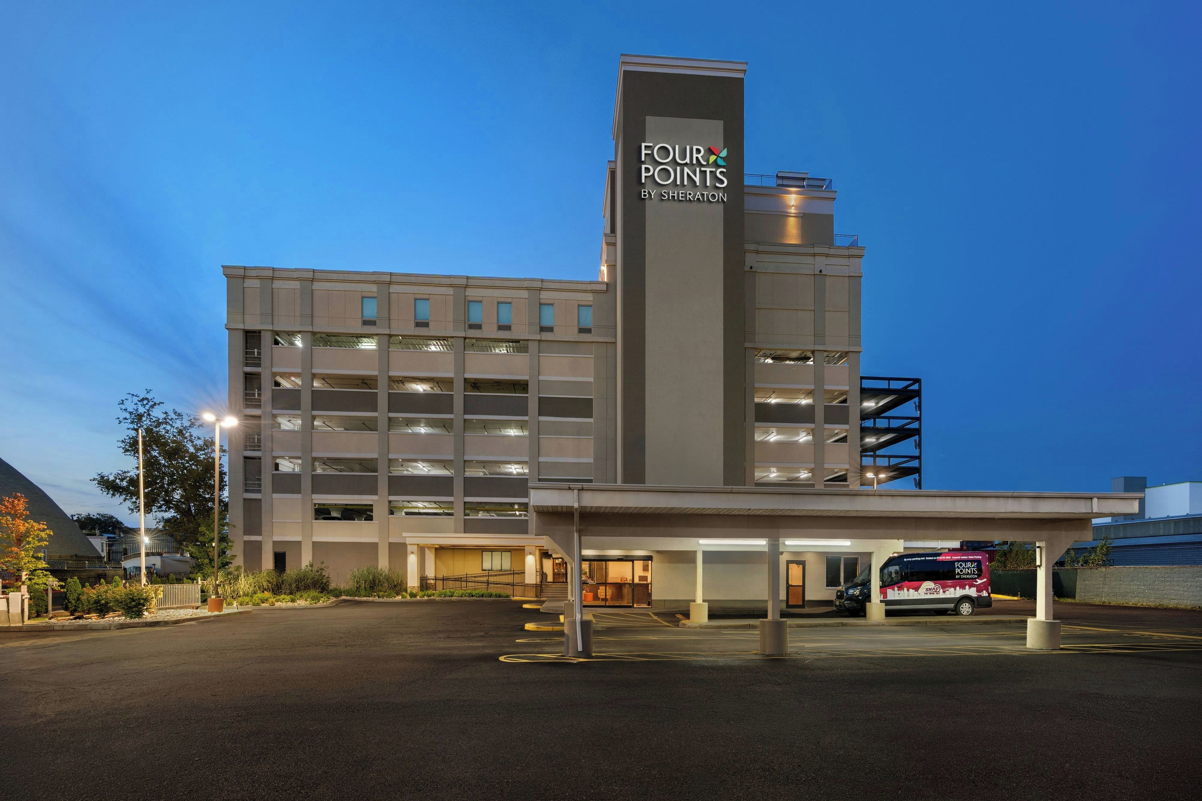 Un hotel de varios pisos con un llamativo letrero de 'FOUR POINTS BY SHERATON', una zona de estacionamiento y servicio de transporte lanzadera.