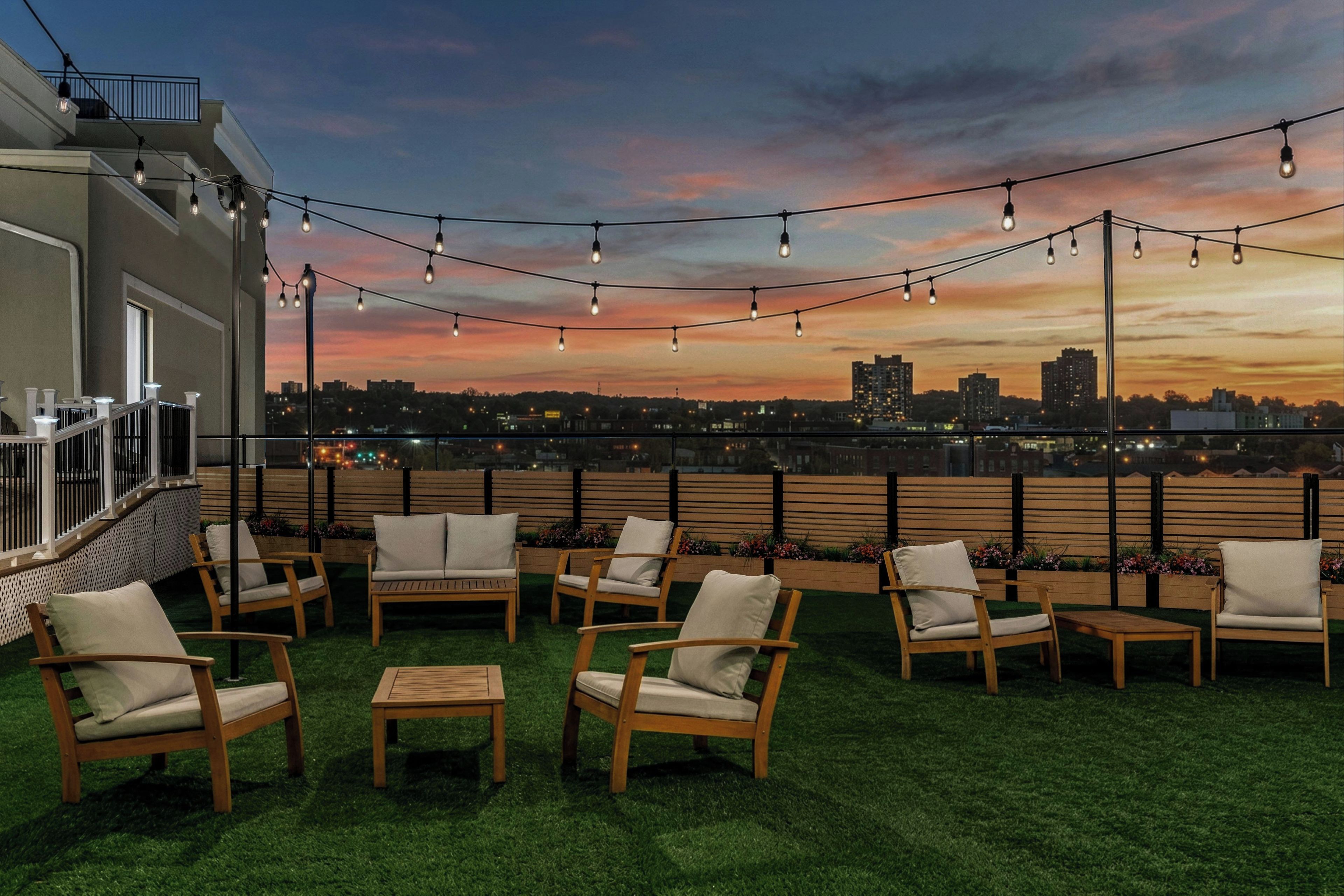 A rooftop terrace with outdoor seating and string lights against a sunset cityscape.
