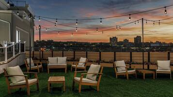 A rooftop terrace with outdoor seating and string lights against a sunset cityscape.