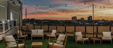 A rooftop terrace with outdoor seating and string lights against a sunset cityscape.