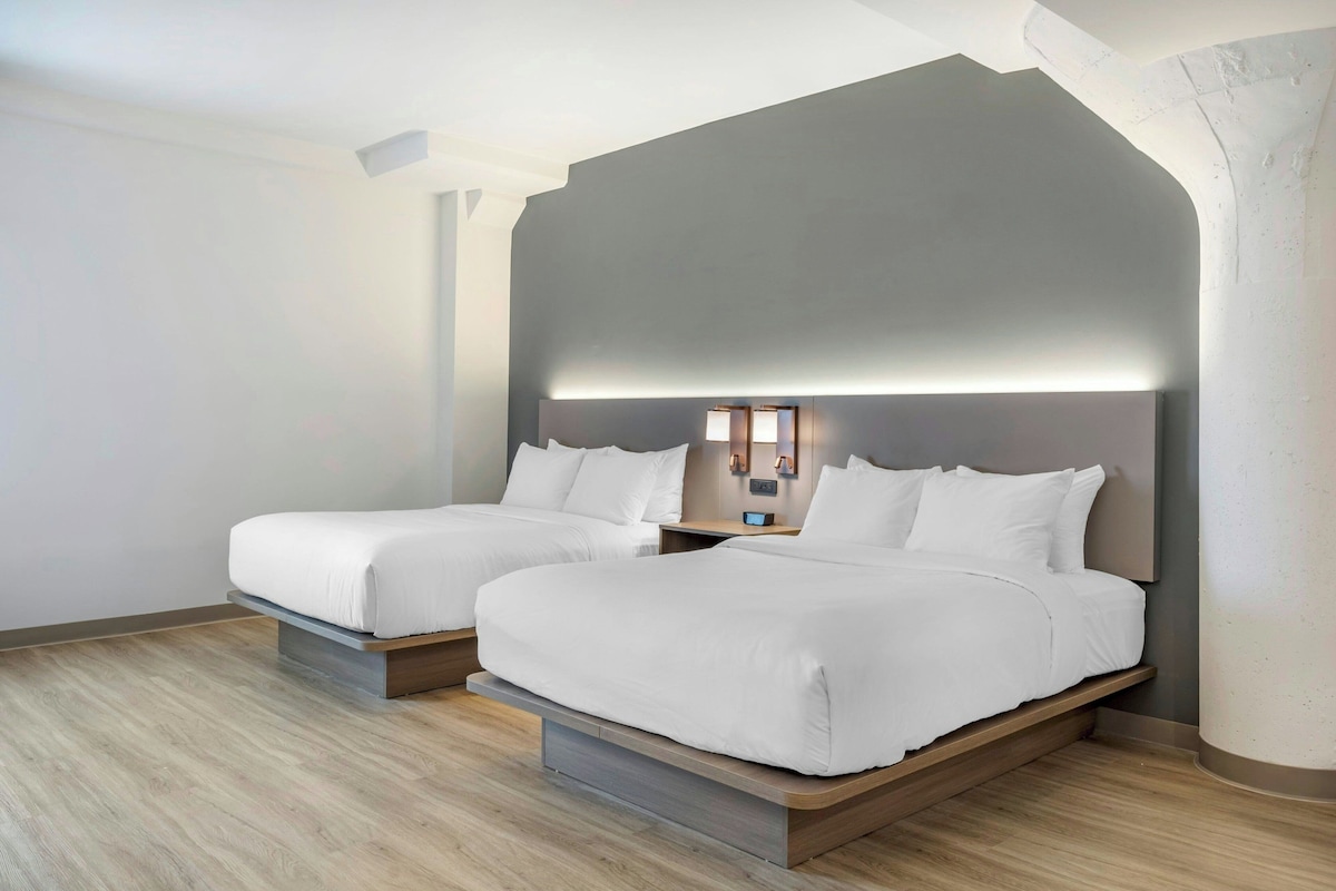 A hotel room with two beds, a modern design, and a nightstand with a lamp.