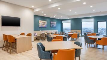 A modern dining area with multiple tables and chairs, a flat-screen TV mounted on the wall, and large windows offering a view of the cityscape.