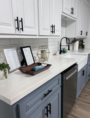A modern kitchen with white cabinets, a dishwasher, and a countertop with a tray and a sign. - Lake Daze Retreat at Kirby ~ Lake Greeson~ (Kirby)