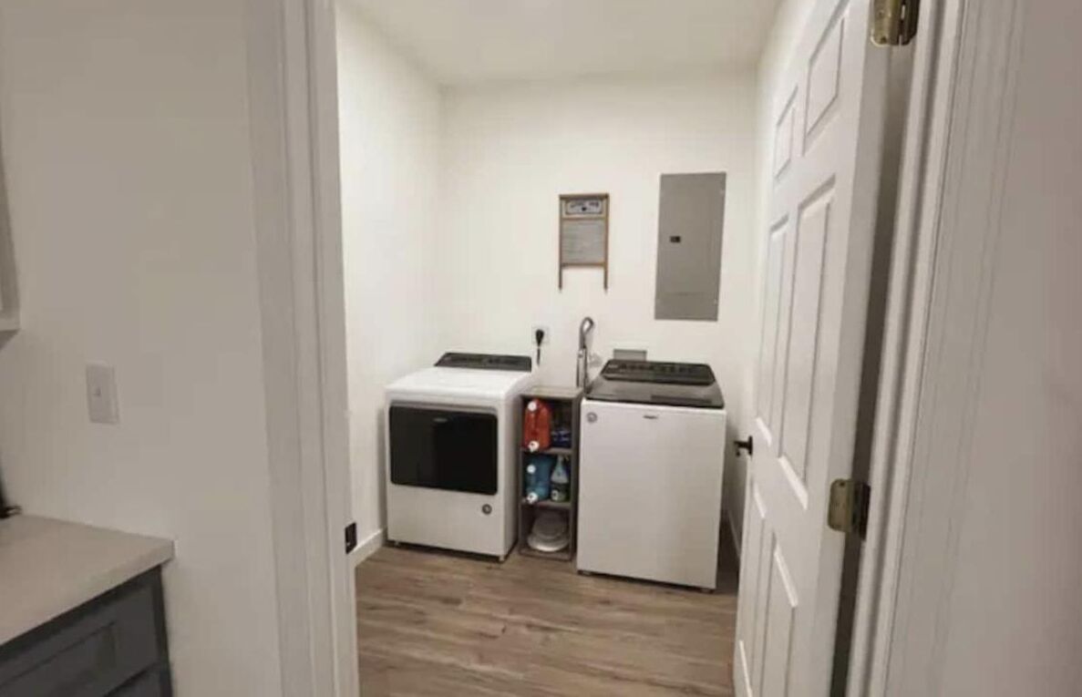 Un lavadero con lavadora y secadora, un estante pequeño con productos de limpieza y una puerta a otra habitación.