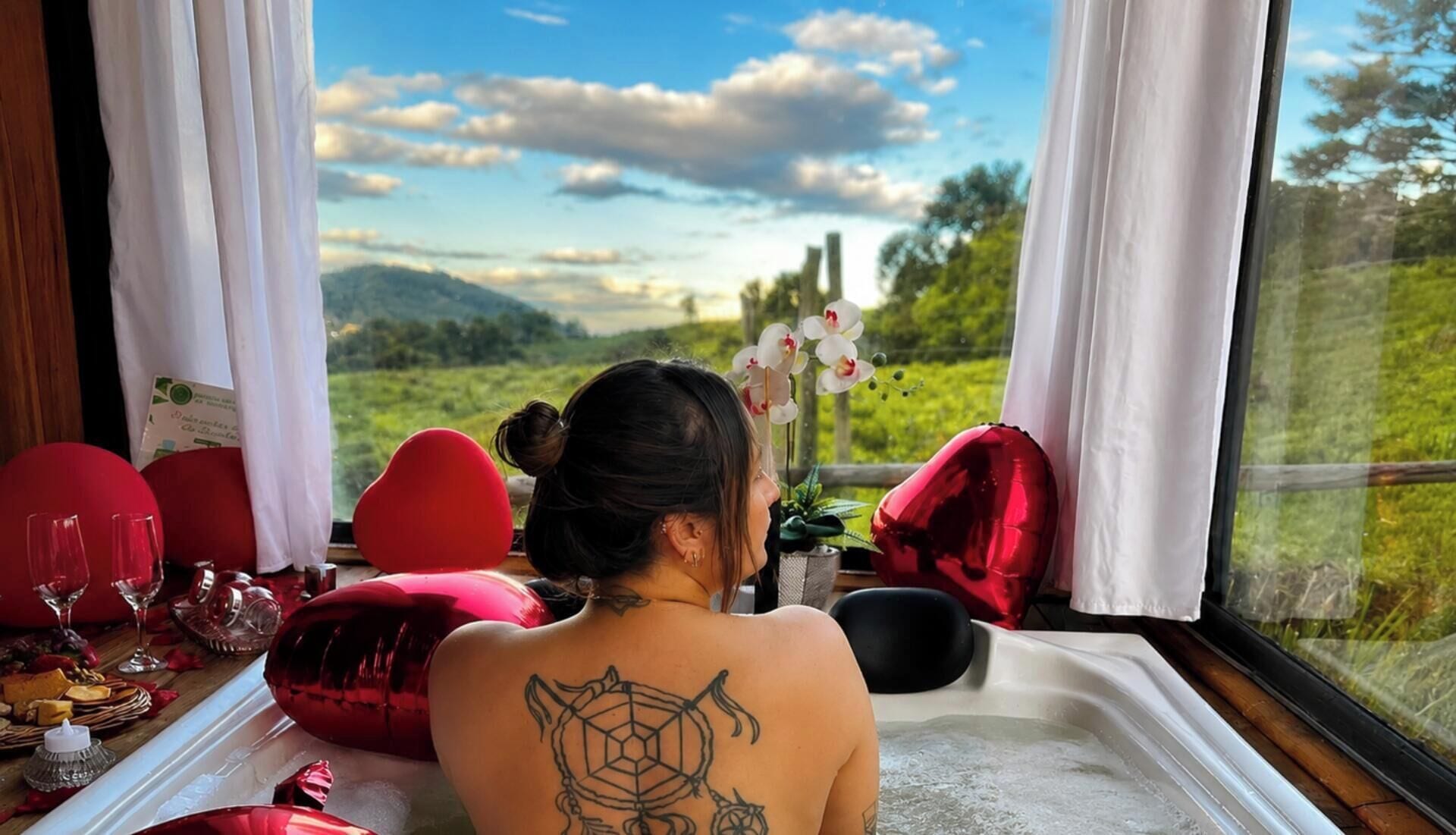 A person seated by a window with a view of a landscape, a table with red cushions, and a tattoo on their back.