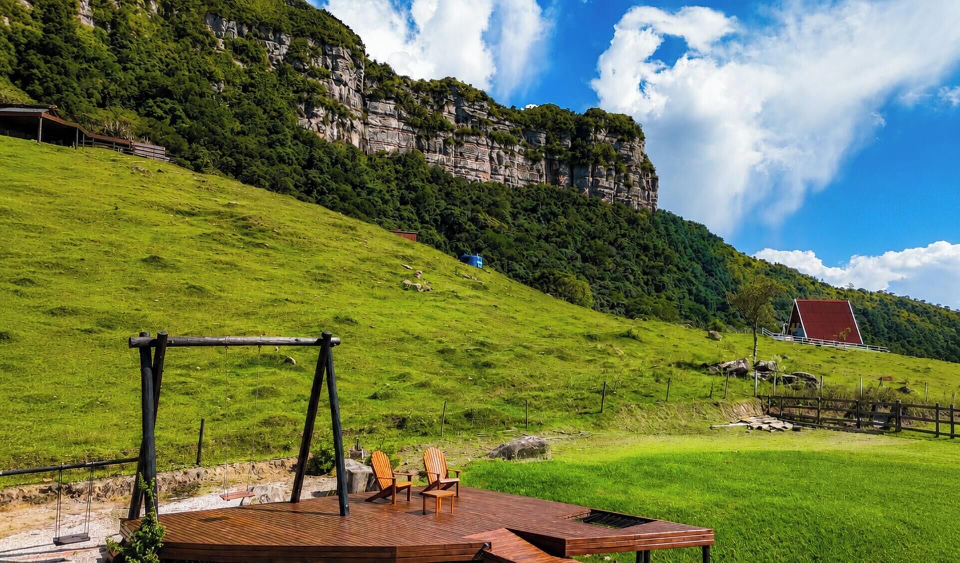 Eine Holzterrasse mit Stühlen und einer Schaukel, mit Blick auf einen grünen Hügel mit einer Klippe und einem roten Gebäude in der Ferne.