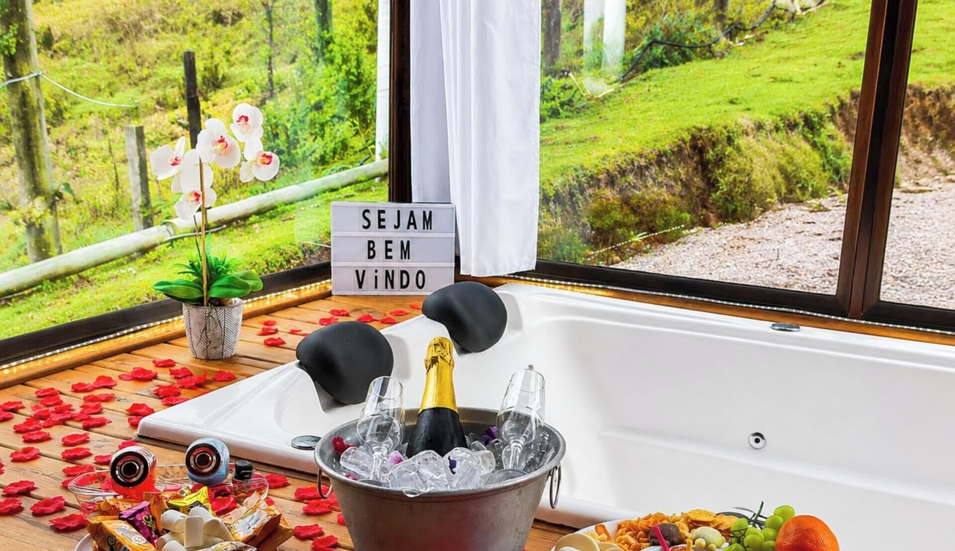 A bathtub with a champagne bottle, ice, and glasses, surrounded by rose petals and a sign that says 'SEJAM BEN VINDO'.