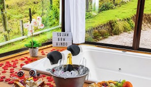 A bathtub with a champagne bottle, ice, and glasses, surrounded by rose petals and a sign that says 'SEJAM BEN VINDO'. - Refúgio Serra da Tartaruga (Alfredo Wagner)