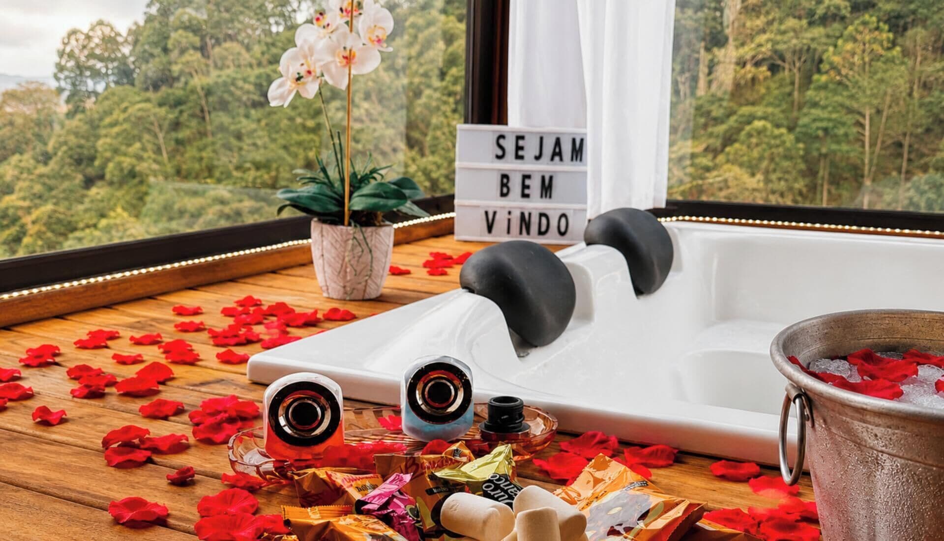 A spa tub with rose petals, a sign reading 'SEJAM BEN VINDO', and a view of a forest through a window.