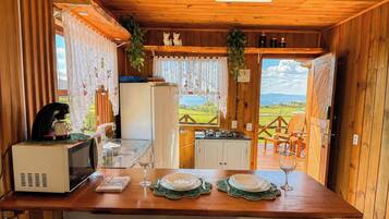 Interior de una cabaña de madera con una mesa de comedor lista para dos personas, una cafetera, un refrigerador y vistas de un paisaje verde a través de una puerta abierta.