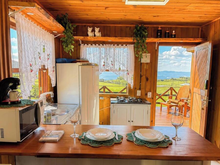 Interior de uma cabana de madeira com uma mesa de jantar posta para dois, uma máquina de café, uma geladeira e vista para uma paisagem verde através de uma porta aberta.