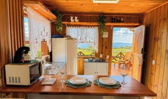 Interior de uma cabana de madeira com uma mesa de jantar posta para dois, uma máquina de café, uma geladeira e vista para uma paisagem verde através de uma porta aberta.
