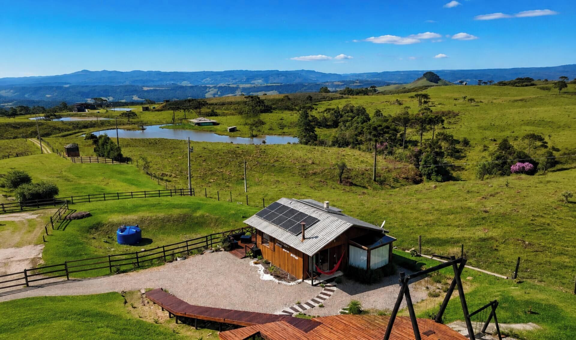 A modern house with a solar panel roof, surrounded by a fenced property with a pond and a hilly landscape in the background.