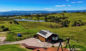 Una casa moderna con techo solar, rodeada por una propiedad cercada, un estanque y un paisaje montañoso al fondo.