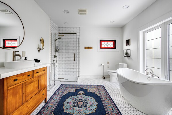 A modern bathroom with a freestanding bathtub, a wooden vanity, a large mirror, and a patterned rug. - Dream Estate Inn (Leamington)