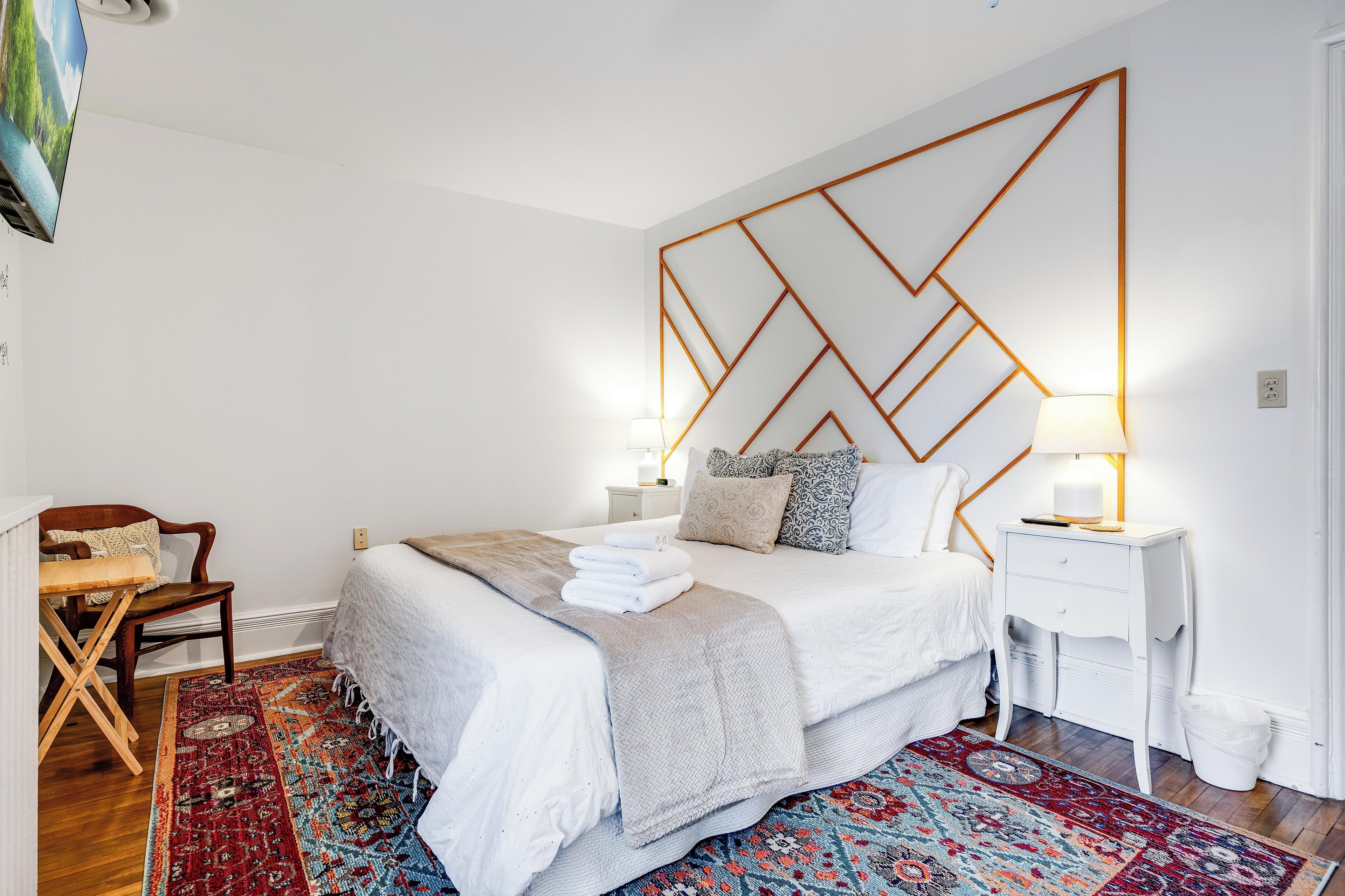 A neatly arranged bedroom with a large bed, a bedside table with a lamp, a chair, and a patterned rug.
