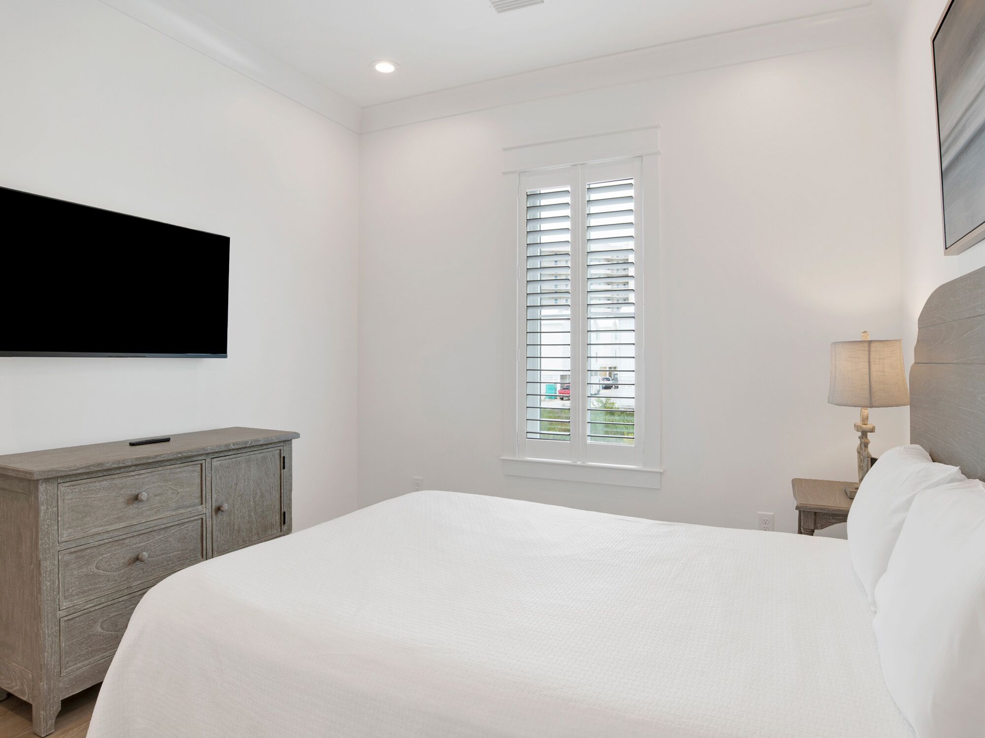 A bedroom with a bed, a dresser, a window with blinds, a lamp, and a TV mounted on the wall.