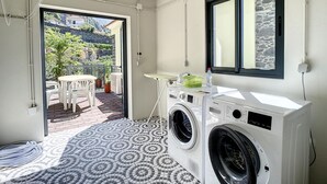 Un área de lavandería con lavadora y secadora, una ventana con pared de piedra, y una puerta que da a un patio exterior con mesa y sillas.