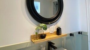 A bathroom with a round mirror, a wooden shelf with a potted plant, a soap dispenser, and a glass door. - PET friendly - Sunny 1 Bedroom Apartment , (Grimsby)