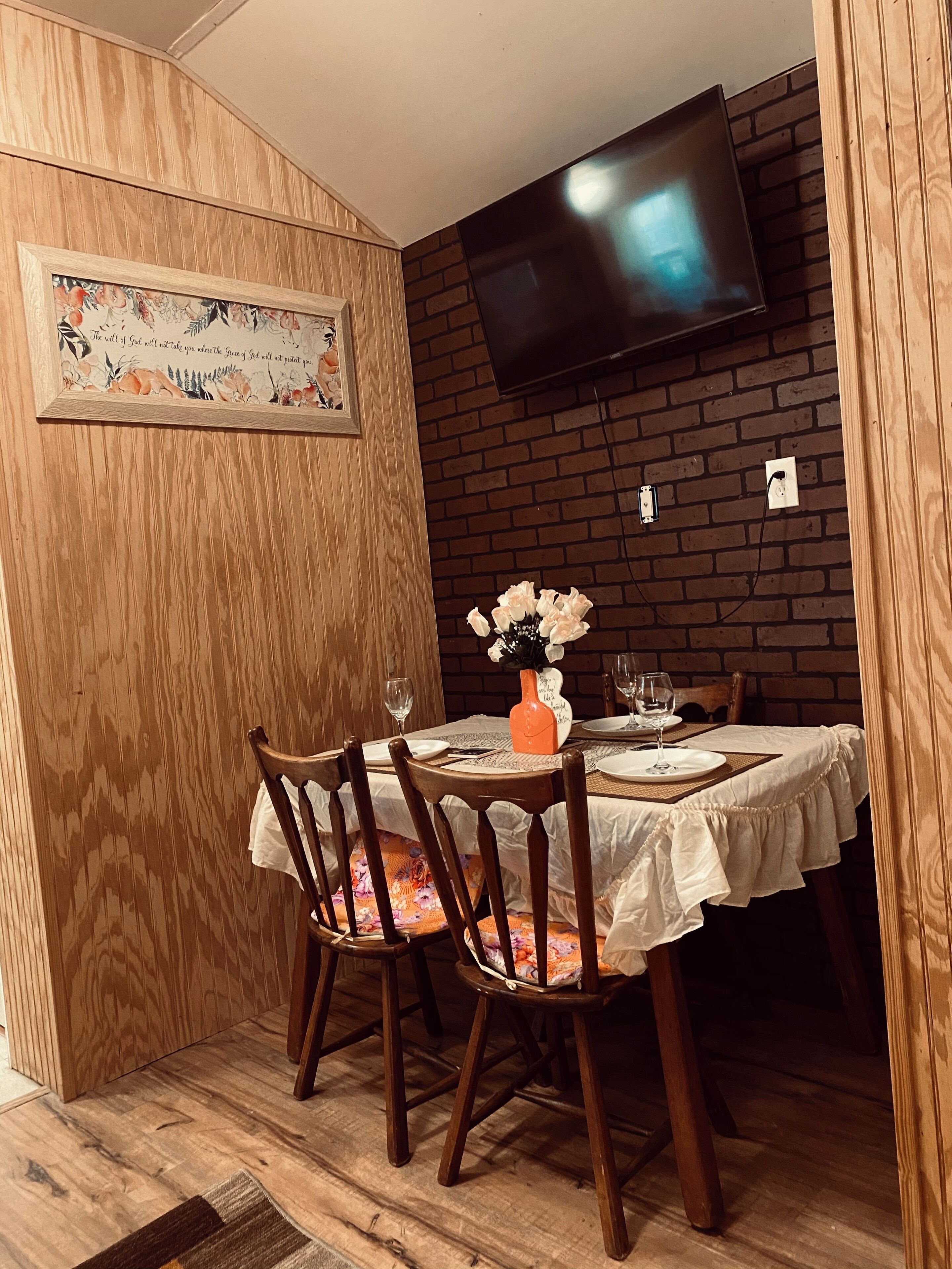 A dining area with a brick wall, a flat-screen TV mounted above the table, and a framed quote on the wall.