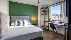 Ein Schlafzimmer mit einer gesteppten Kopfteil, einem Bett mit weißer Bettwäsche, einem Schreibtisch mit Stuhl und Blick auf Gebäude durch ein Fenster.