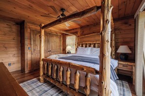 A wooden cabin-style bedroom with a four-poster bed, a ceiling fan, and a nightstand with a lamp.