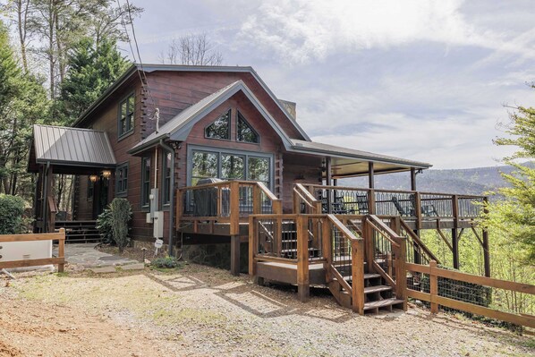 A log cabin with a covered porch, a wooden staircase, and a deck overlooking a forested area. - Above the Clouds: Summer Mountain Cabin with Hot Tub & Fireplace (Blue Ridge)
