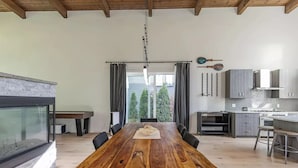 A spacious dining area with a wooden table and chairs, a kitchen with modern appliances, and a view of trees through large windows. - Large Modern Home in Squamish (Squamish)