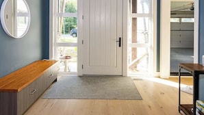 A room with a high ceiling, a white door with glass panels, a wooden bench, a round mirror, and a small table with a box. - Large Modern Home in Squamish (Squamish)