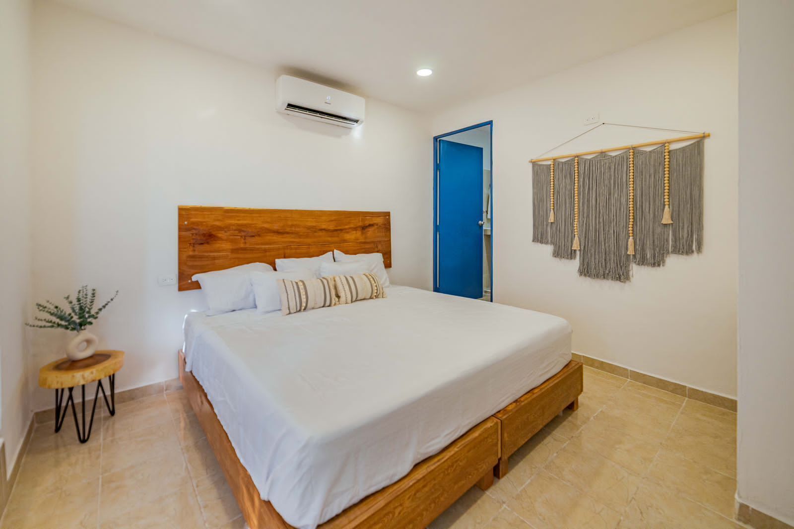 Een slaapkamer met een houten bed, wit beddengoed, een airconditioningapparaat, een blauwe deur, een kleine plant en een macramé wanddecoratie.