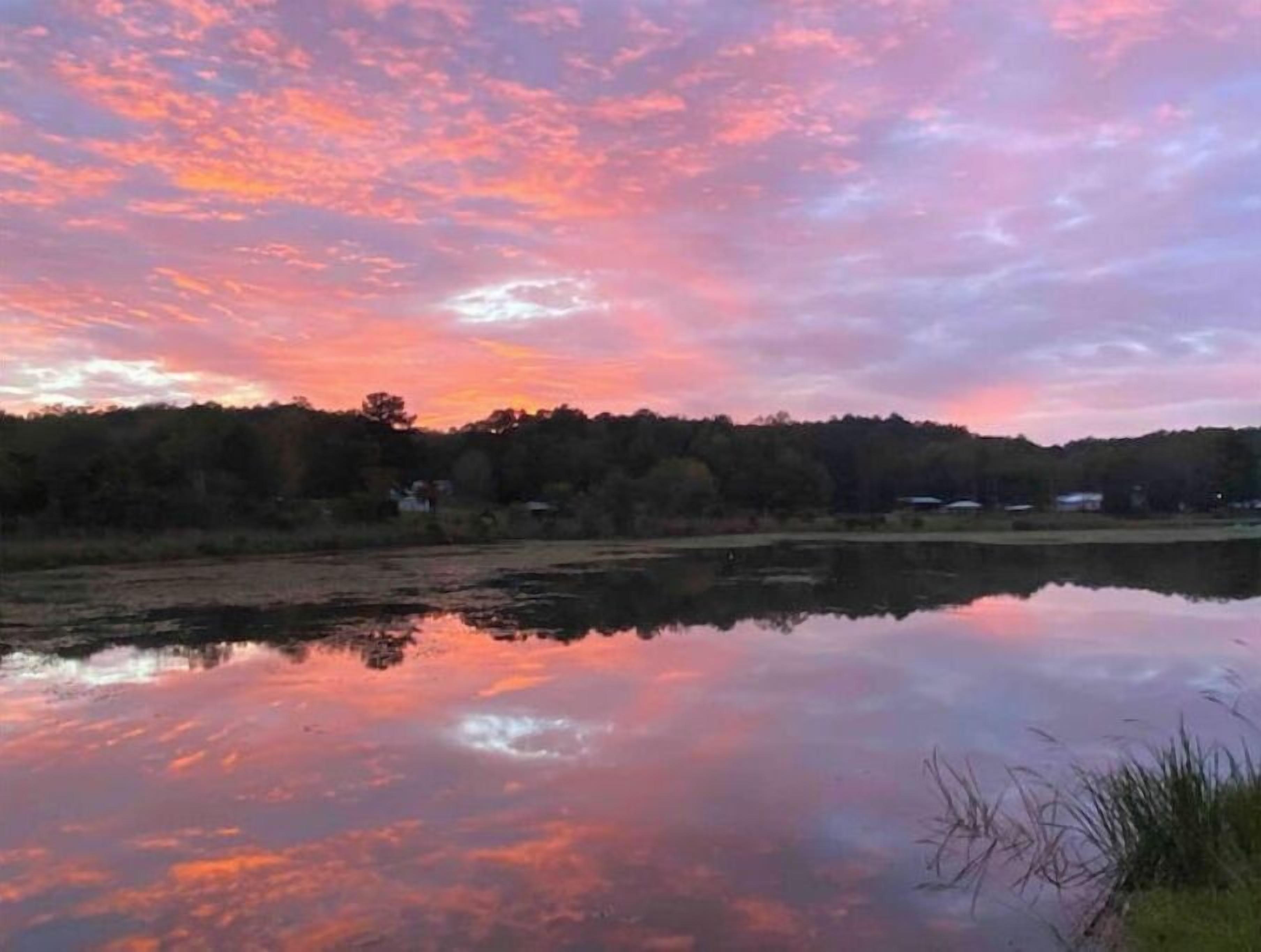 A calm lake reflecting a sunset sky with trees and buildings in the background.