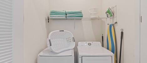 Un espace buanderie comprenant deux machines à laver blanches, un panier à linge blanc, une étagère avec des serviettes pliées et un porte-mop.