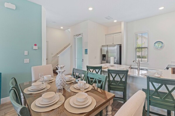 A dining area with a wooden table set for four, surrounded by green chairs. A kitchen with a stainless steel refrigerator and a sink is visible in the background. - 8000 Lost Shaker Lane (Kissimmee)