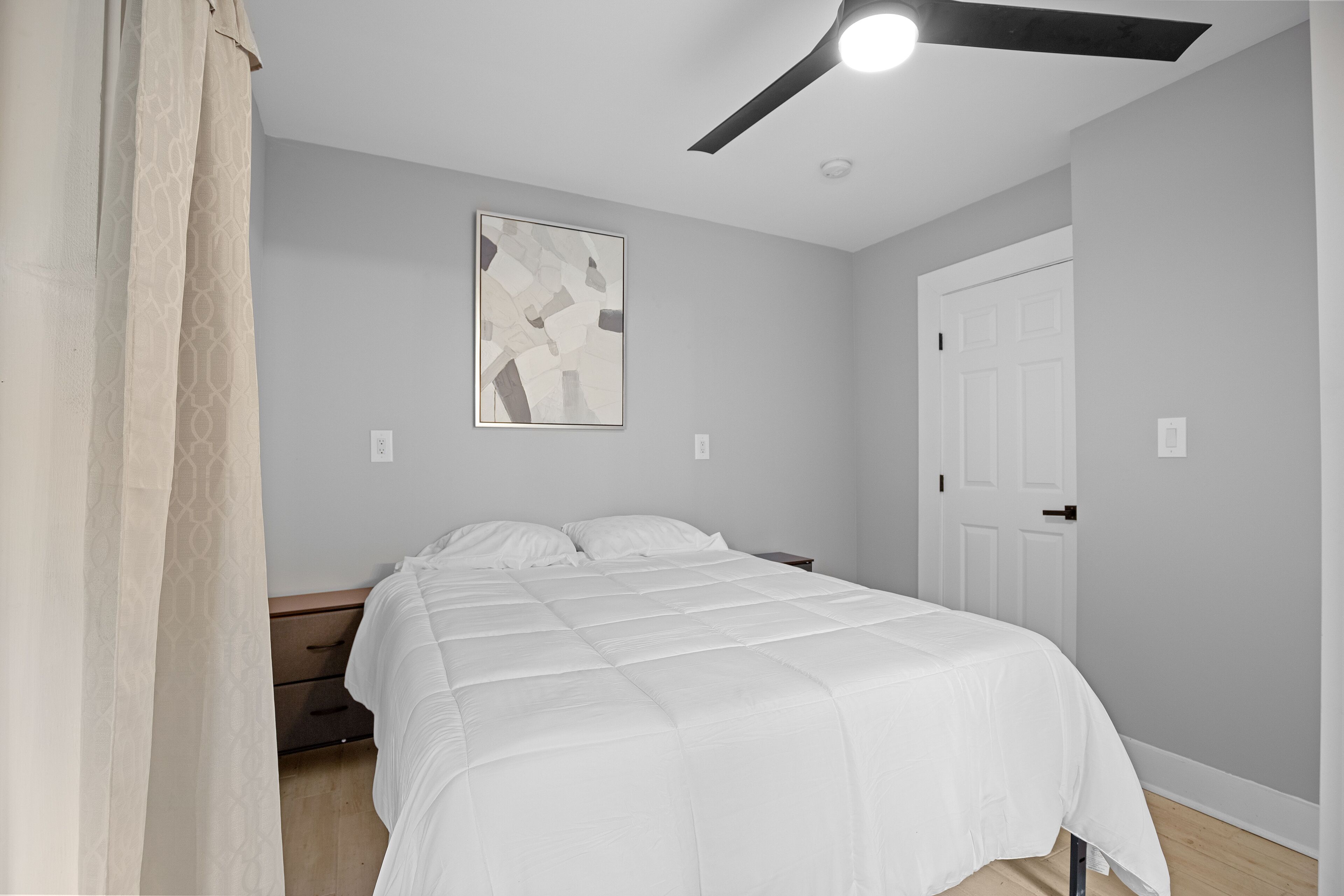 A bedroom with a bed, a ceiling fan, a door, and a wall-mounted abstract artwork.