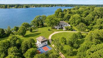 A large house with a tennis court, surrounded by dense trees and a lake.