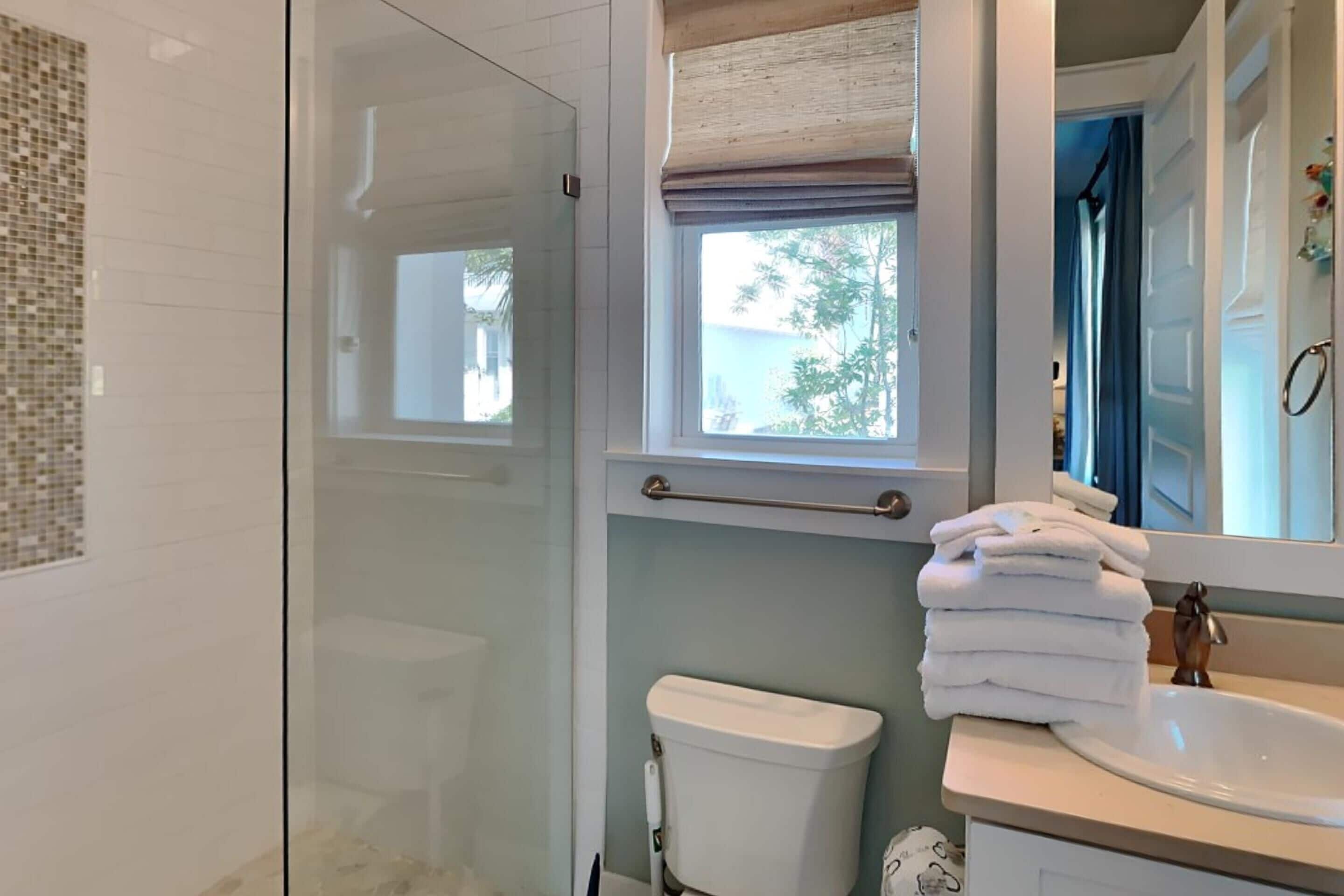A bathroom with a glass shower enclosure, a toilet, a sink with a mirror, and a window with a view of trees.