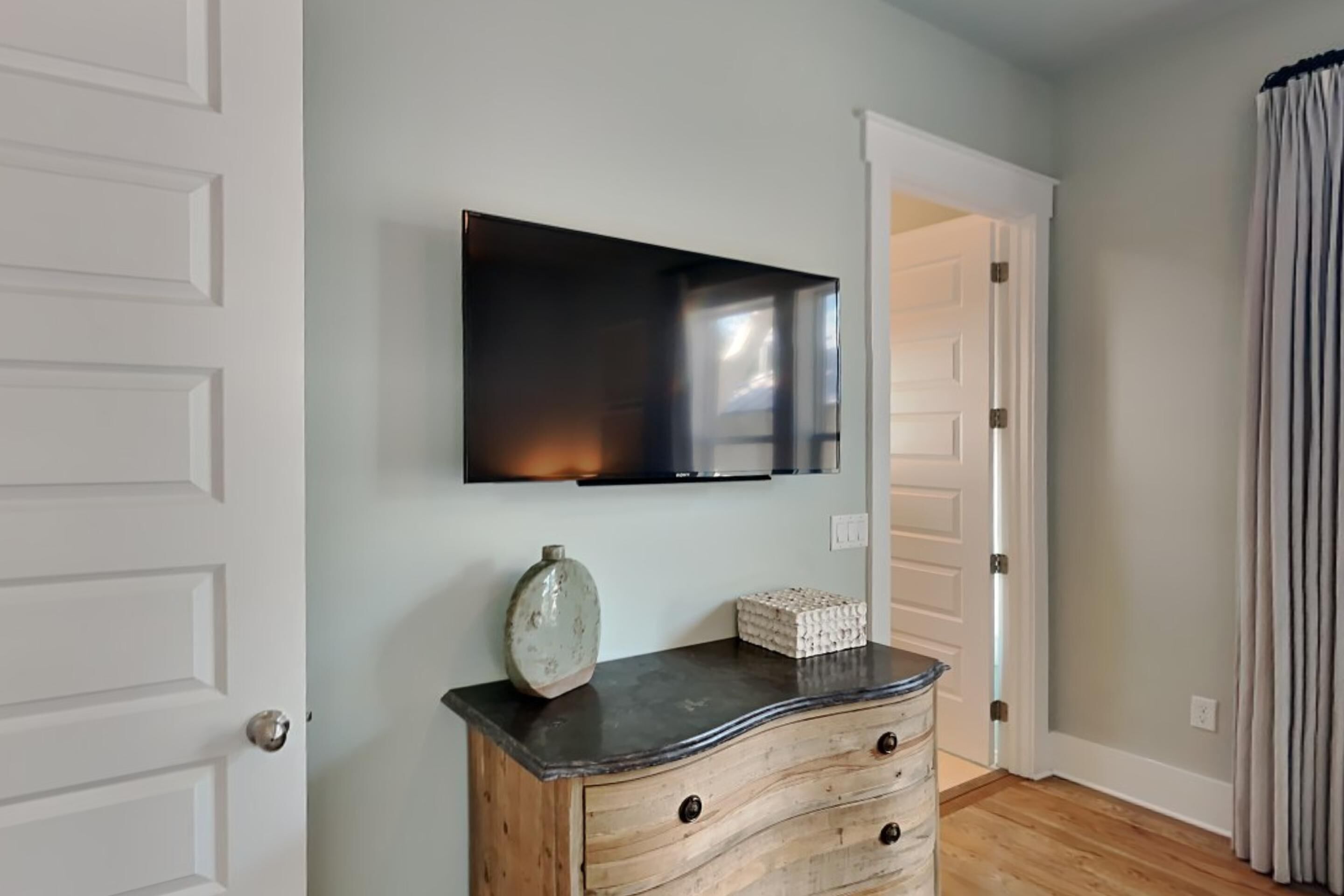 A room with a flat-screen TV mounted on the wall, a wooden dresser with drawers, a decorative vase, and a door leading to another room.